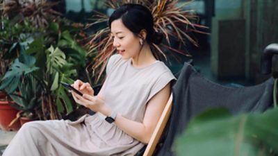 Getty 1312631510 Beautiful young Asian woman using smartphone while relaxing on deck chair in the backyard, surrounded by beautiful houseplants. Lifestyle and technology