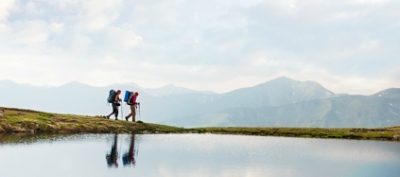 Two hikers passing by a calm mountain lake reflecting the sky